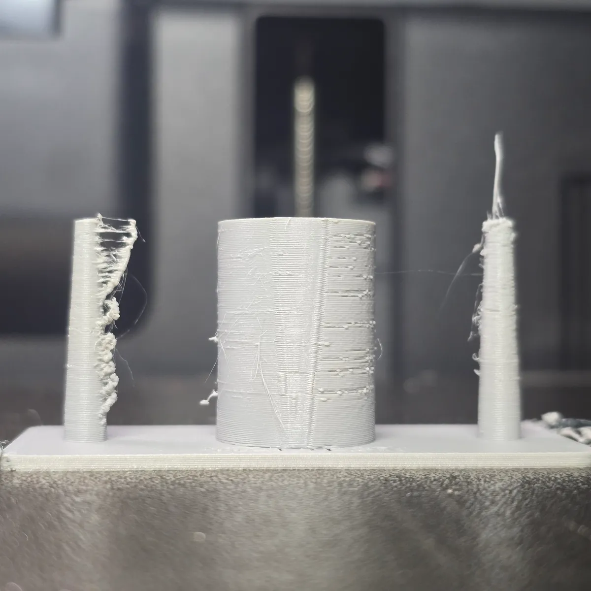 Print 1 in grey PETG: the left post wrapped in a band of hairs and blobs, rows of small zits on the ring, a long whisker on the right post and a thin string from the ring to it.